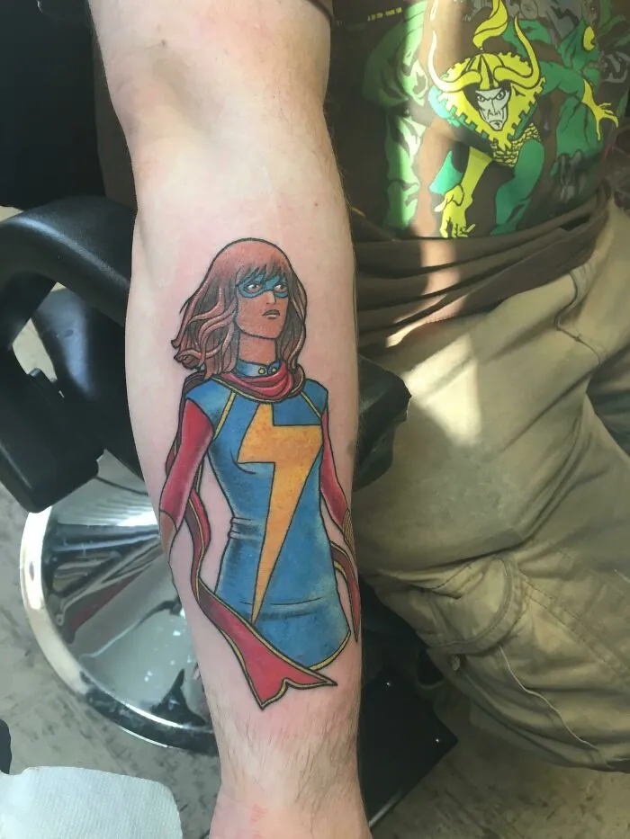 Colorful Marvel tattoo featuring Ms. Marvel on a forearm, showcasing comic art style with vibrant blue and yellow costume.