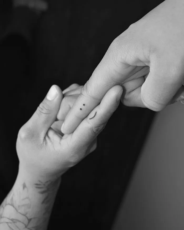 Minimalist tattoo of a tiny face on a finger, held by another hand.