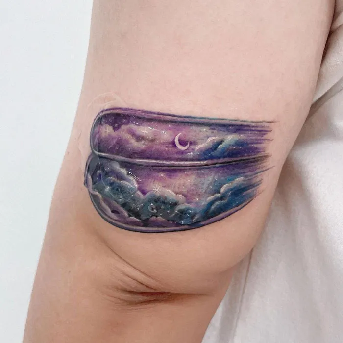 Watercolor tattoo of a night sky with clouds and a crescent moon on an arm.