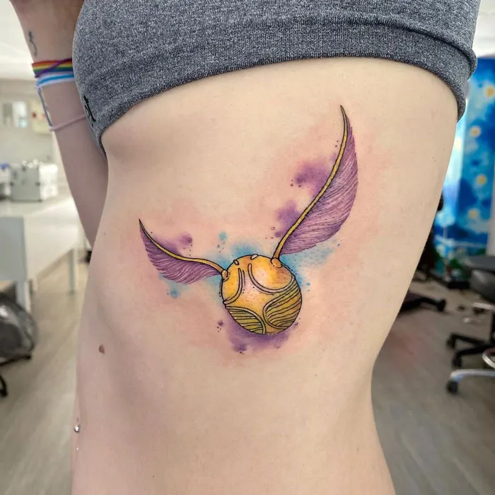 Colorful watercolor tattoo of a golden snitch with wings on a person's side.