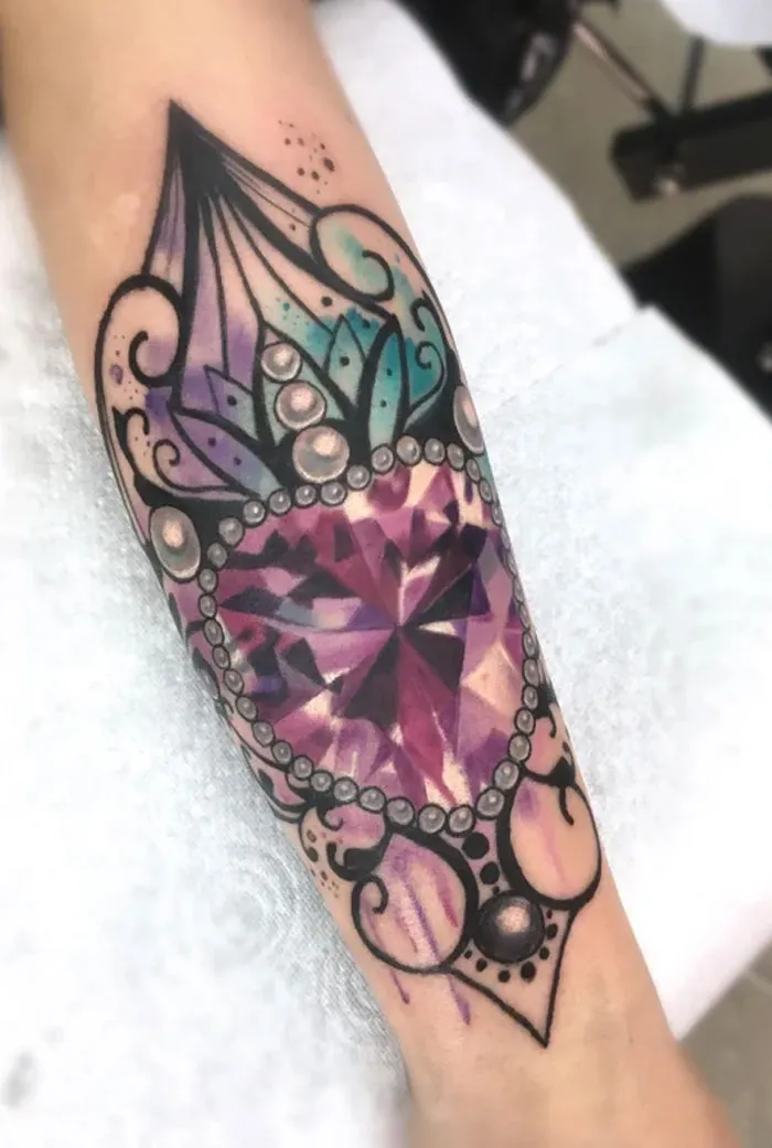 Watercolor tattoo with a purple gem and ornate design on forearm.
