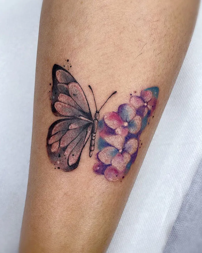 A beautiful watercolor tattoo of a butterfly with floral elements on a forearm.