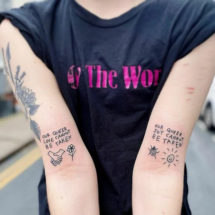 Tattoos on forearms reading "Our queer love cannot be taken" and "Our queer joy cannot be taken," with simple line art.