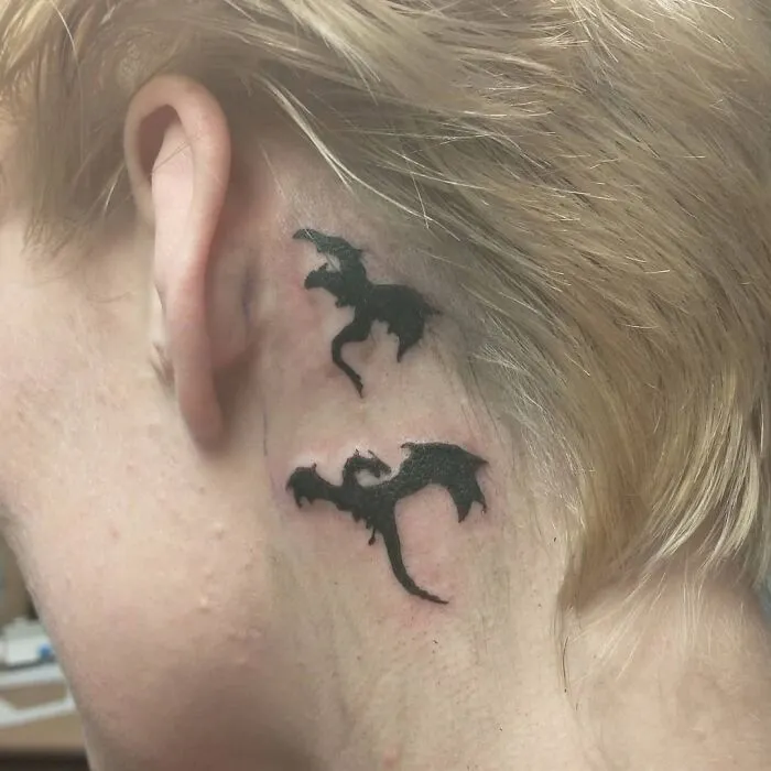 Ear tattoo of two black dragons behind the ear against blonde hair.