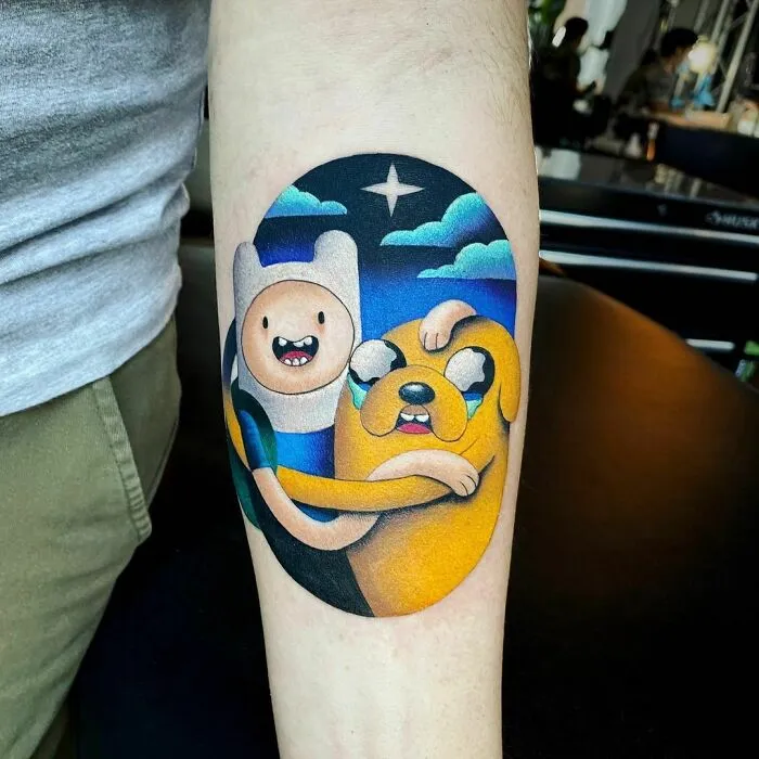 Finn and Jake cartoon tattoo, vibrant colors, on forearm for a nostalgic touch.