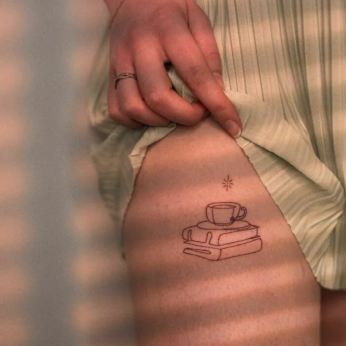 Minimalist tattoo of a coffee cup and books on a person's thigh, enhancing aesthetic appeal with simple design.