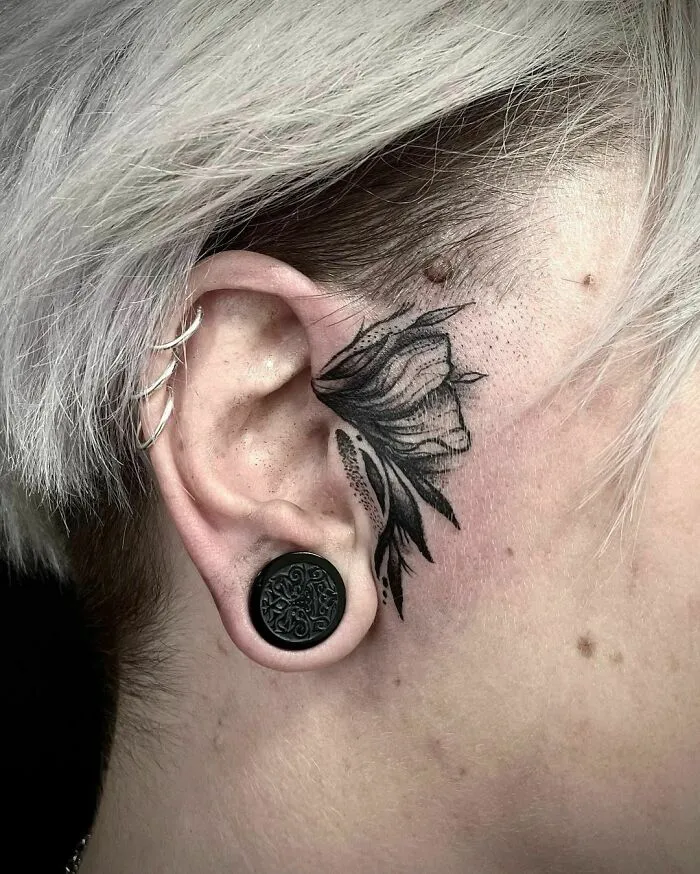 Ear tattoo with floral design, black gauge earring, and silver helix piercings in blonde hair.