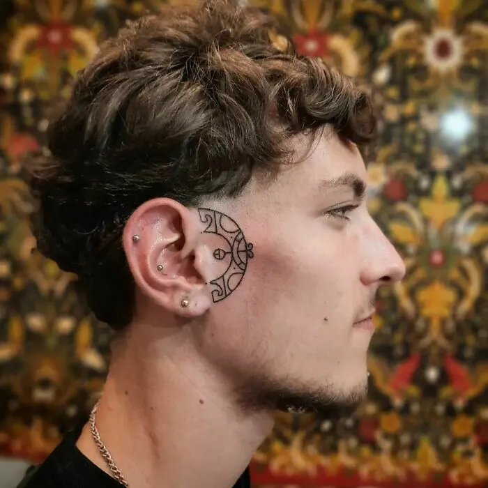 Ear tattoo featuring a geometric design behind the ear with multiple piercings, set against a decorative background.