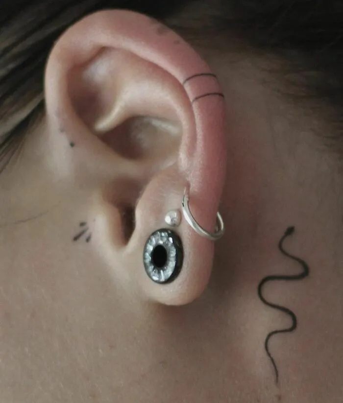 Close-up of an ear featuring minimalist tattoos and piercings, including a hoop and a spiral design.