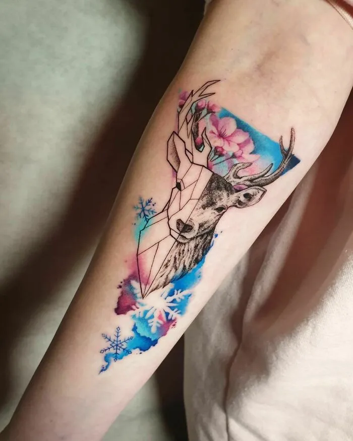 Watercolor tattoo of a deer with geometric design, pink flowers, and blue snowflakes on forearm.