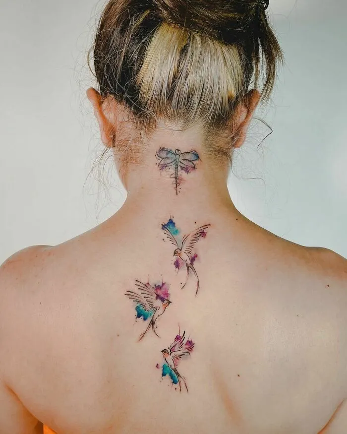 A woman's back features beautiful watercolor tattoos of birds and a dragonfly, showcasing vibrant ink inspiration.