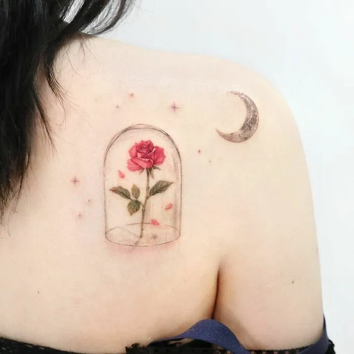 Watercolor tattoo of a rose under a glass dome with a crescent moon on a shoulder, showcasing inkspiration.