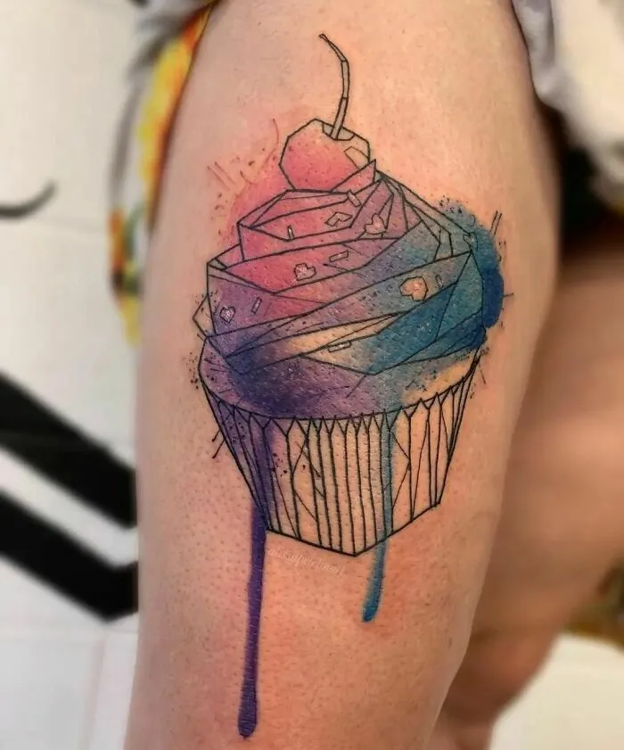 Watercolor tattoo of a cupcake with geometric shapes and vibrant colors on a person's arm.