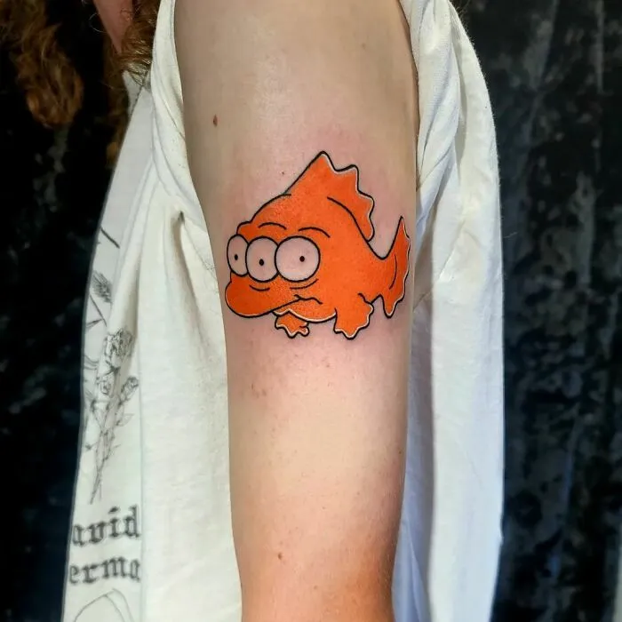Cartoon tattoo of a three-eyed orange fish on a person's arm, inspired by animated shows.