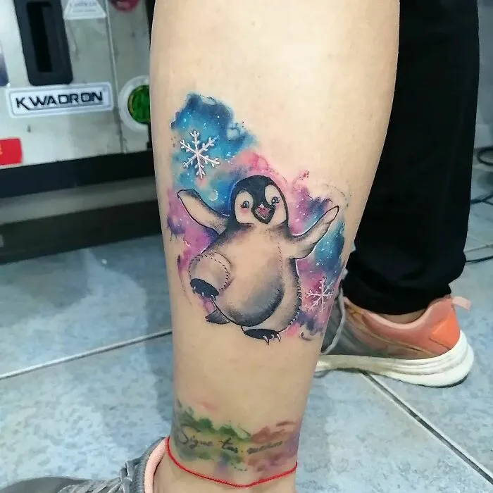 Colorful watercolor penguin tattoo on a leg, featuring snowflakes and vibrant ink.