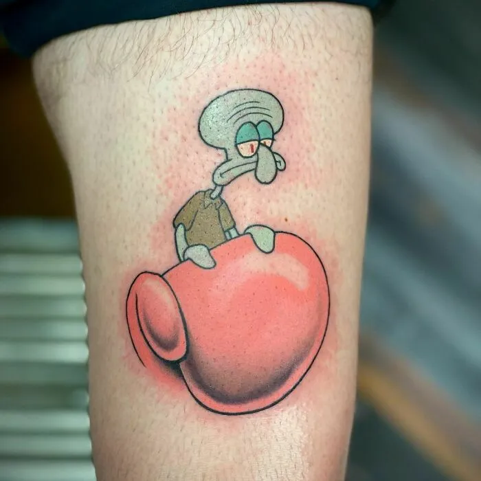 Cartoon tattoo featuring a grumpy character sitting on a large pink object, inspired by animated shows.