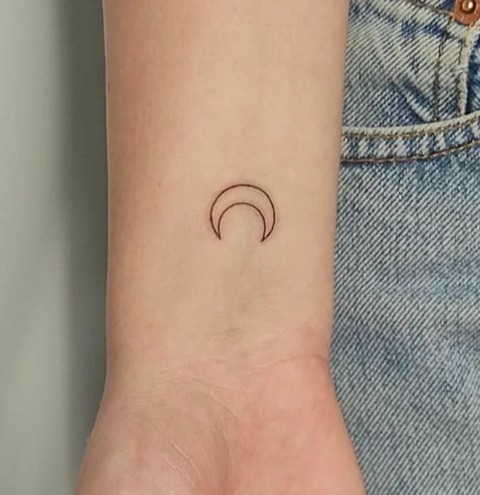 Minimalist tattoo of a crescent moon on a forearm, set against denim fabric.