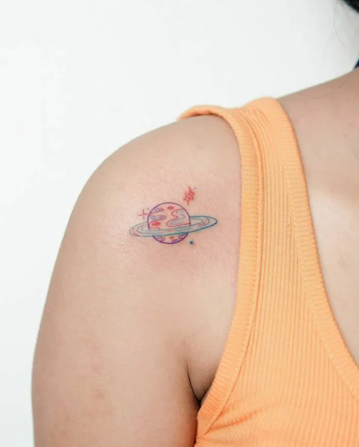 Minimalist tattoo of a colorful planet with a ring on a person's upper arm, showcasing aesthetic simplicity.