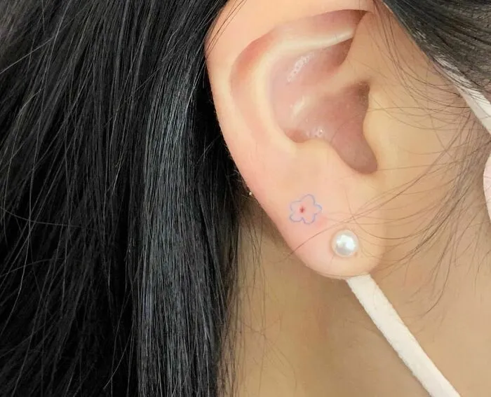 Minimalist flower tattoo near ear with pearl earring, showcasing simple yet aesthetically pleasing design.