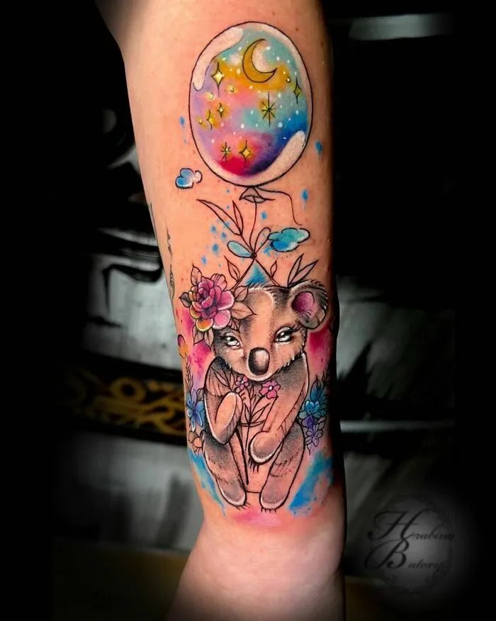 Watercolor tattoo of a koala with flowers and a moon balloon in vibrant colors on an arm.