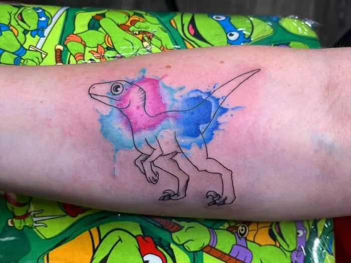 Watercolor tattoo of a dinosaur in blue and pink hues on a forearm, set against a colorful background.