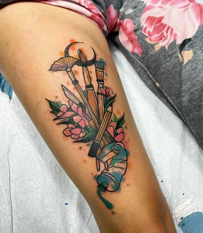 Watercolor tattoo of art supplies and flowers on an arm, featuring vibrant colors and intricate details.