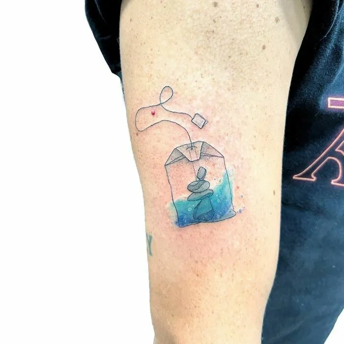 Watercolor tattoo of a tea bag with water and stones, inked on a person's arm.