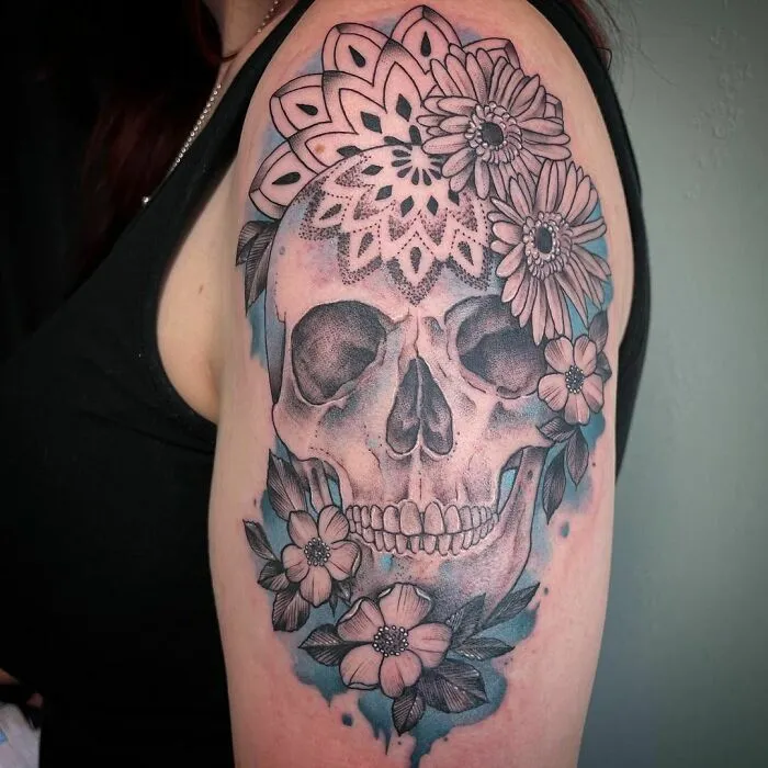 Watercolor tattoo of a skull adorned with floral and mandala designs on an arm.