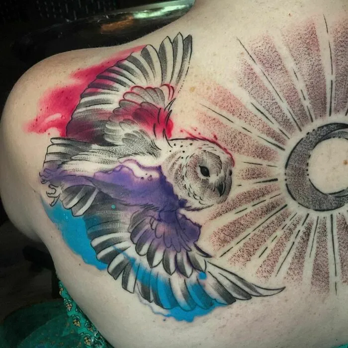 Watercolor tattoo of an owl in flight on shoulder, featuring vibrant colors and a sunburst pattern.