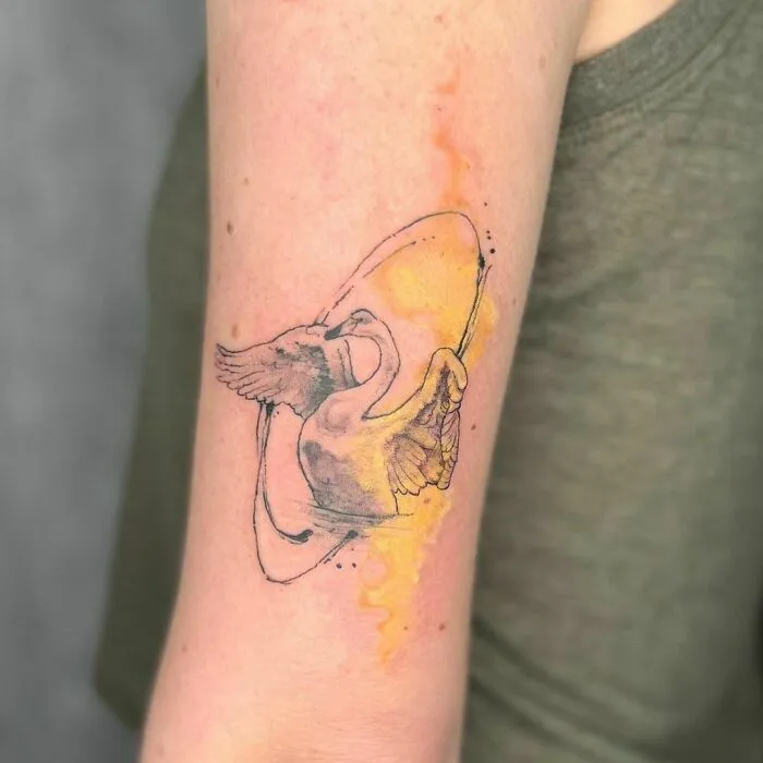 Watercolor tattoo of a swan with wings spread, surrounded by a splash of yellow and orange ink on a person's arm.