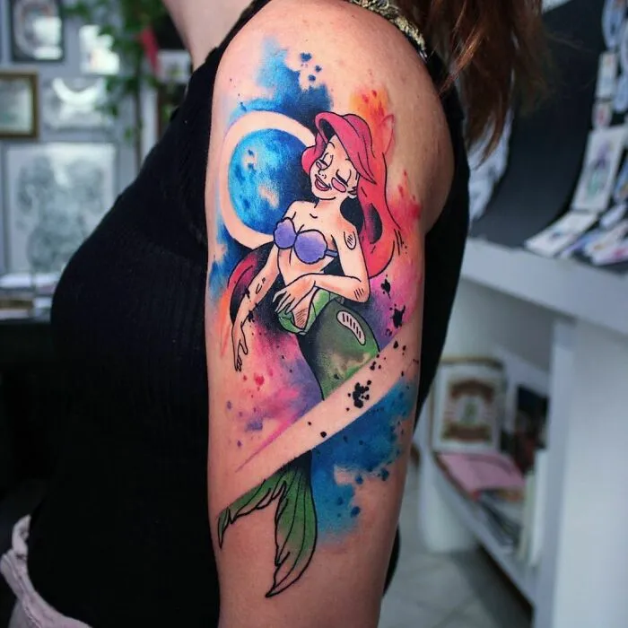 Watercolor tattoo of a mermaid in vibrant blues and reds on a person's upper arm.