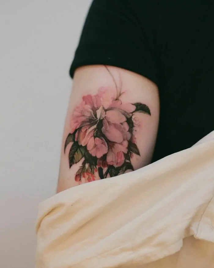 Pink floral watercolor tattoo on arm, featuring delicate petals and green leaves, showcased on a light-skinned person.