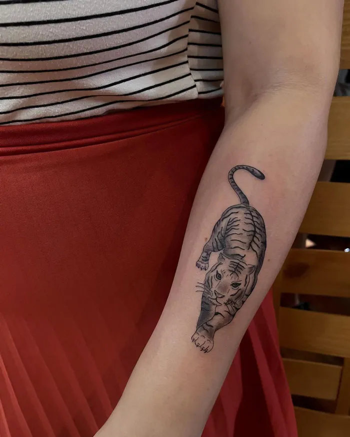 A tiger tattoo on forearm, showcasing animal kingdom art, worn by a person in a striped top and red skirt.