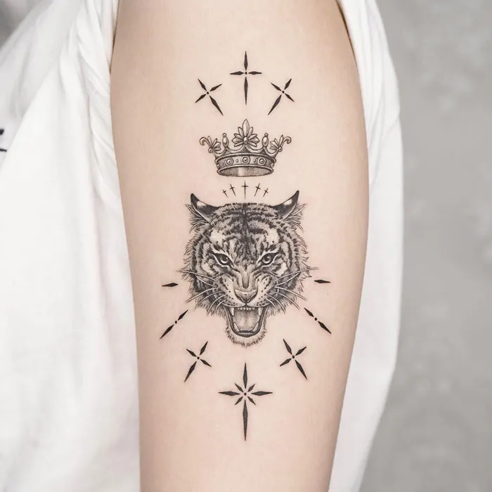Animal tattoo of a roaring tiger with a crown on an arm, surrounded by star-like patterns.