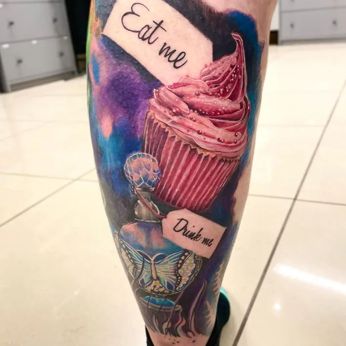 Lower leg tattoo of a colorful cupcake with "Eat me" and "Drink me" labels, featuring a butterfly design.
