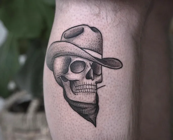 Cowboy skull calf tattoo, featuring a detailed design with a hat and bandana on the leg.