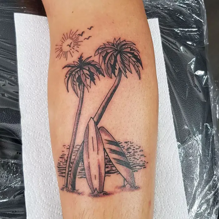 Lower leg tattoo featuring palm trees and surfboards on a beach under the sun.