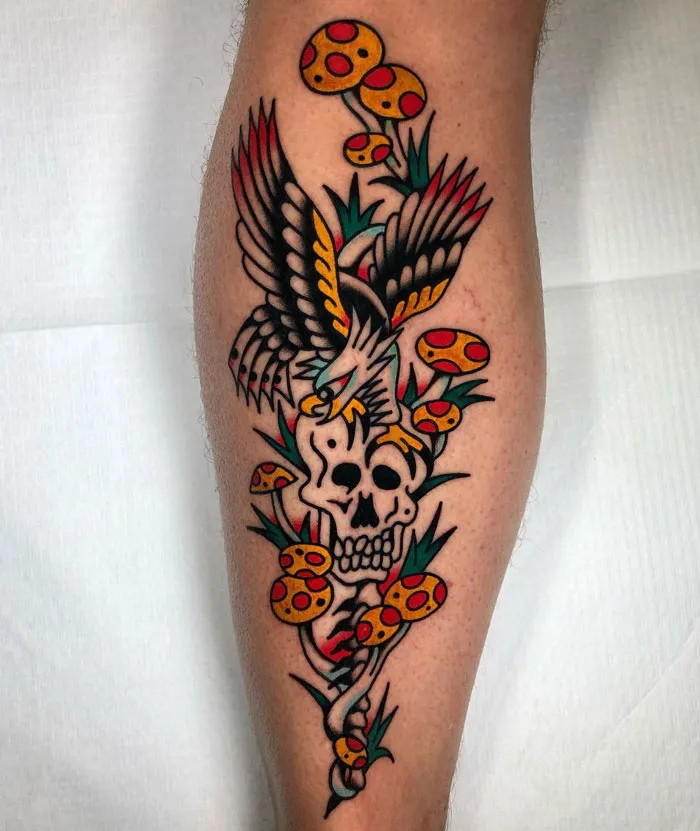 Colorful lower leg tattoo featuring a skull, eagle, and mushrooms.