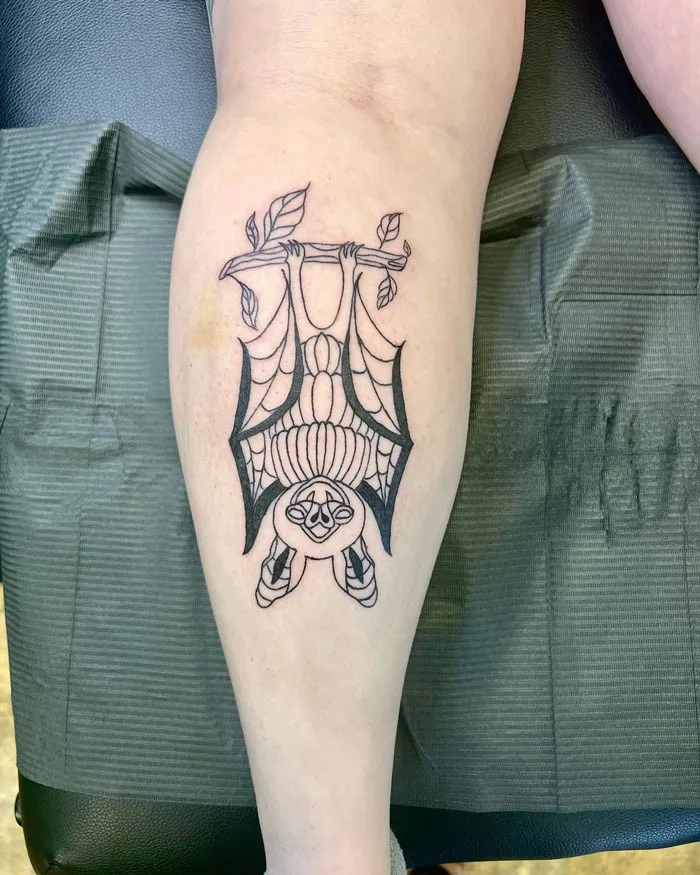 Lower leg tattoo of a stylized bat hanging upside down with leaves on a branch.
