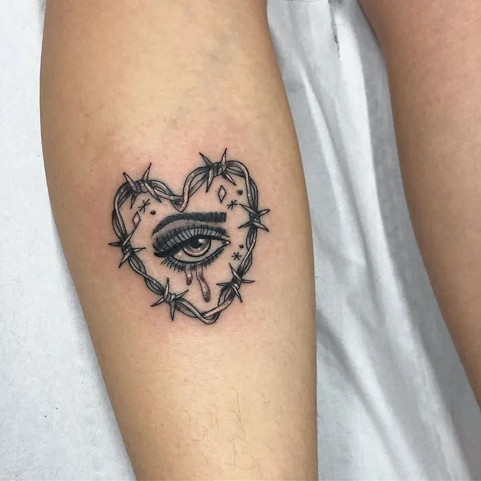 Heart-shaped eye tattoo with barbed wire on lower leg.