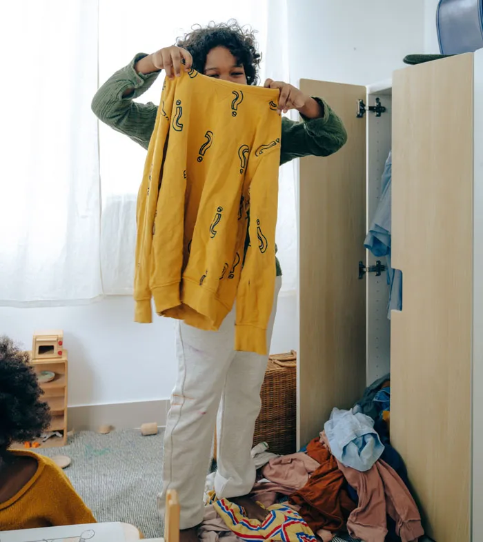 Child holding a yellow shirt with question marks, organizing clothes at home to avoid boredom.