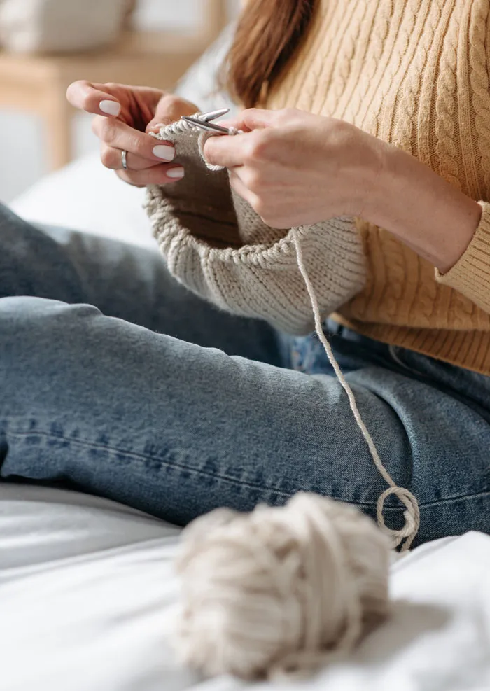 Woman knitting at home, wearing a cozy sweater and jeans, with yarn and needles, engaging in a fun activity to avoid boredom.