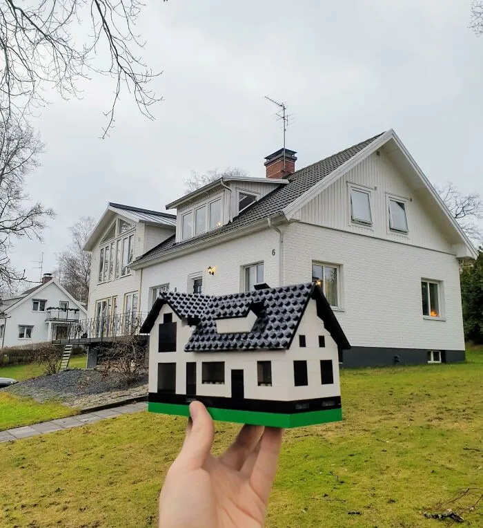 Hand holding a detailed LEGO house build in front of a real house, showcasing incredible LEGO builds as works of art.