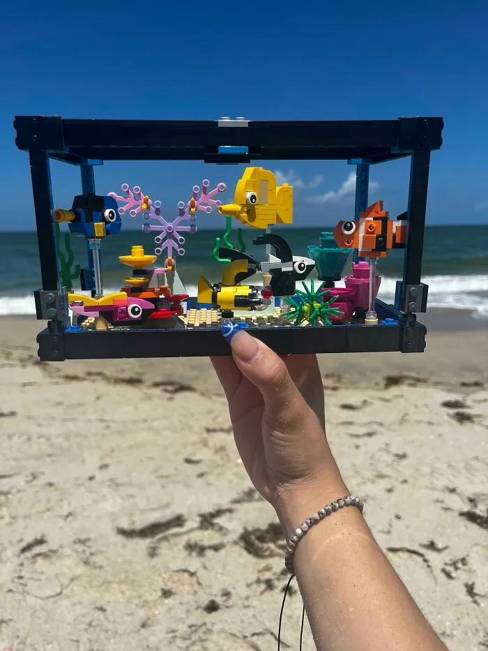 Hand holding colorful LEGO builds of fish and underwater scene with ocean and beach in the background.