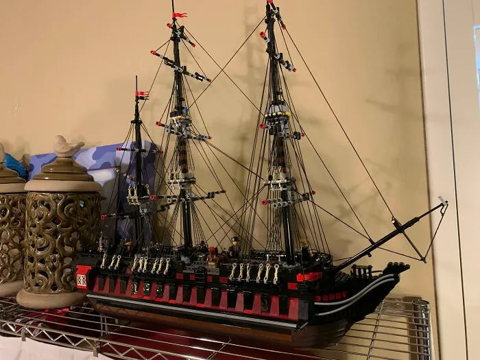 Intricate LEGO builds of a detailed tall ship with multiple masts and rigging displayed on a metal shelf indoors.