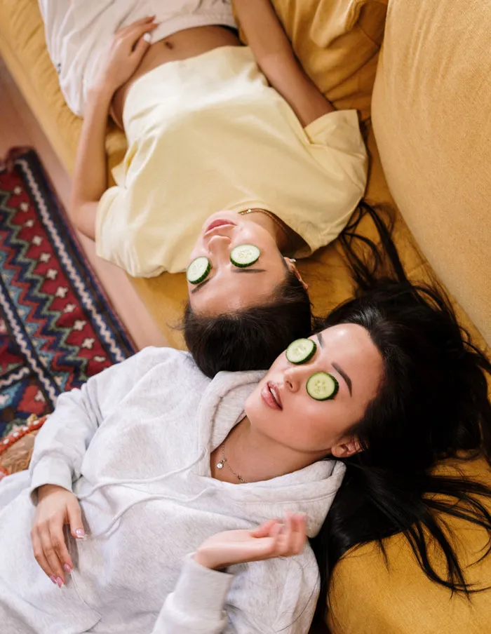 Two women relaxing at home with cucumber slices on eyes, lying on a mustard couch.