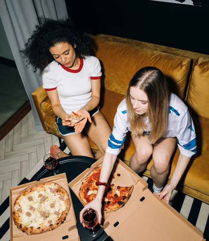 Two women enjoying pizza and wine at home, having fun together on a cozy evening.