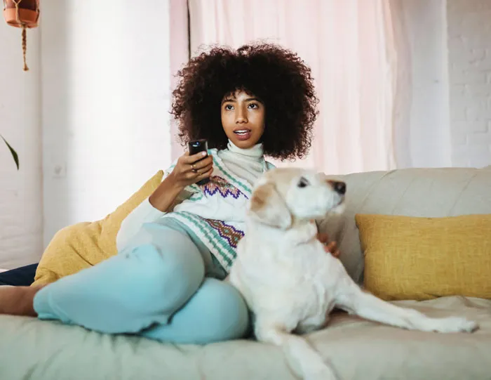 A woman at home relaxing on a couch with her dog, avoiding boredom with a remote in hand.