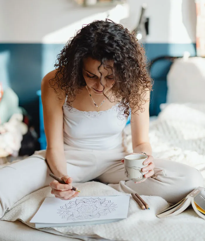 A woman doing a fun coloring activity at home, sitting cross-legged and holding a mug, avoiding boredom.