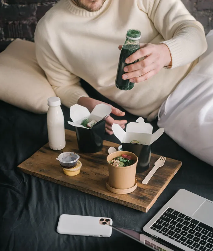 Man enjoying a meal in bed with takeout containers, drinks, and a laptop; fun things to do at home.